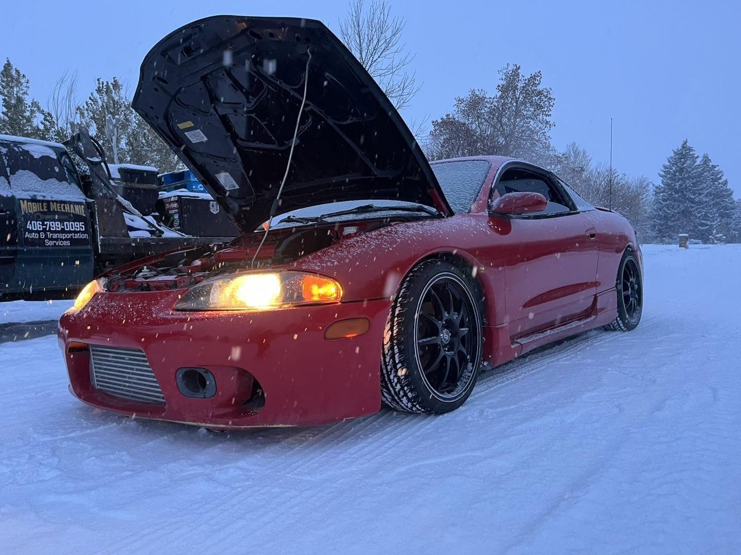 Customer's red sports car with the hood up getting repaired in falling snow, 406 Mobile Mechanic work truck parked behind